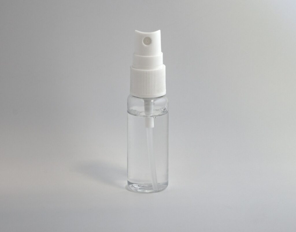 Een klein, doorzichtig plastic spuitflesje met een witte spuitmond en dop, gevuld met een transparante vloeistof, rechtopstaand tegen een effen, lichtgrijze achtergrond.