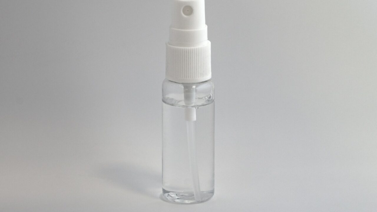 Een klein, doorzichtig plastic spuitflesje met een witte spuitmond en dop, gevuld met een transparante vloeistof, rechtopstaand tegen een effen, lichtgrijze achtergrond.