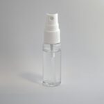 Een klein, doorzichtig plastic spuitflesje met een witte spuitmond en dop, gevuld met een transparante vloeistof, rechtopstaand tegen een effen, lichtgrijze achtergrond.