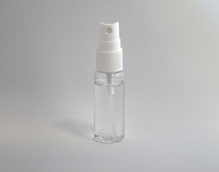 Een klein, doorzichtig plastic spuitflesje met een witte spuitmond en dop, gevuld met een transparante vloeistof, rechtopstaand tegen een effen, lichtgrijze achtergrond.